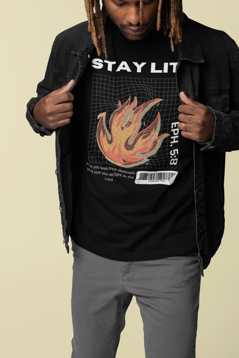 A man wearing a black Christian graphic t-shirt with a flame design and the text 'STAY LIT' and 'EP. 5:8', layered under an open black denim jacket, paired with gray pants, against a solid beige background.
