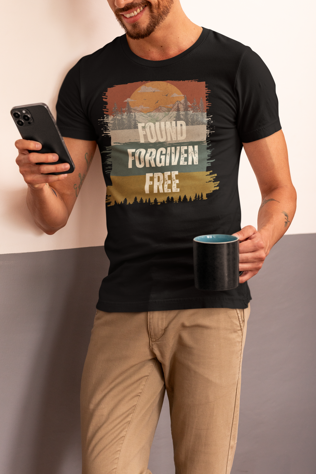 A man wearing a black Christian graphic t-shirt with a retro sunset design and the text 'FOUND FORGIVEN FREE', paired with khaki pants, holding a phone and a mug.