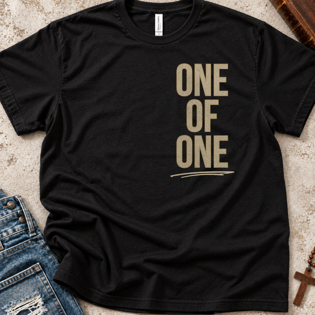 A black short-sleeve t-shirt with the words 'ONE OF ONE' printed vertically in a muted tan serif font on the left chest, styled in a flat lay with blue jeans and a wooden cross nearby.