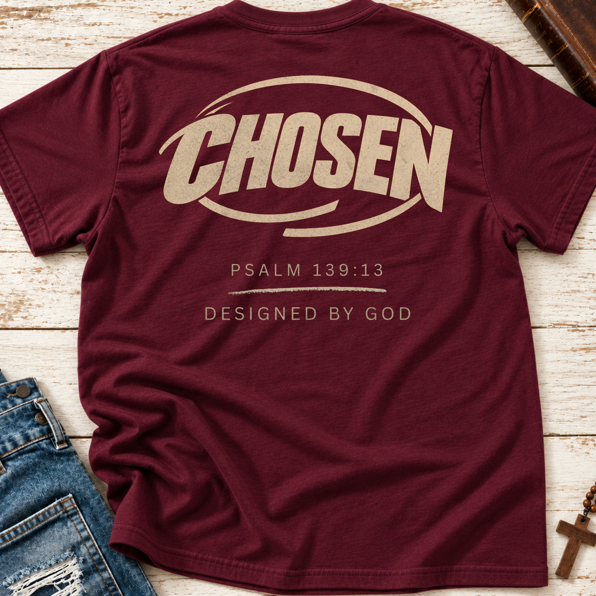 A maroon short-sleeve t-shirt laid flat on a whitewashed wood surface, featuring a large beige graphic on the back that reads 'CHOSEN' with 'PSALM 139:13' and 'DESIGNED BY GOD' below. Blue jeans and small decorative props are partially visible at the edges.