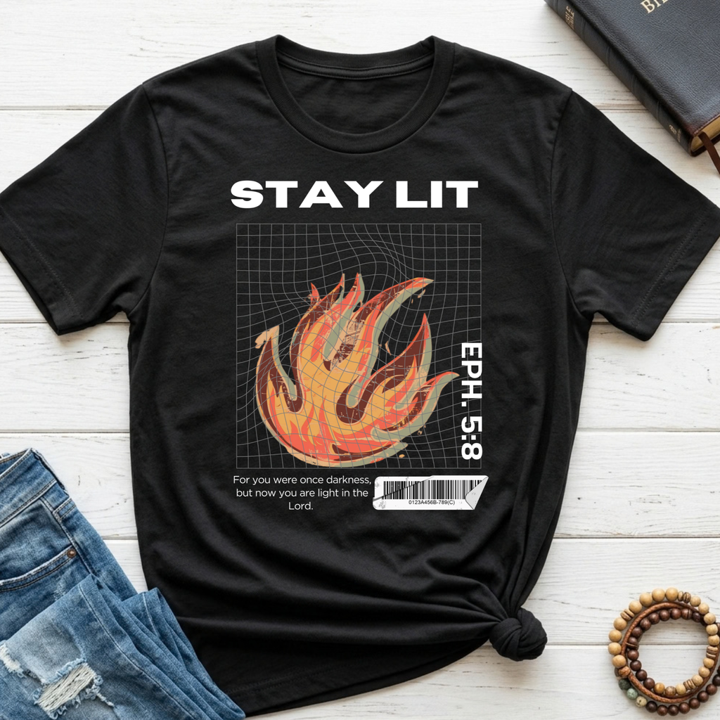 A black graphic t-shirt laid flat on a white wood background, styled with blue jeans, a book, and a beaded bracelet. The shirt features a white 'STAY LIT' text and a vertical 'EPH.