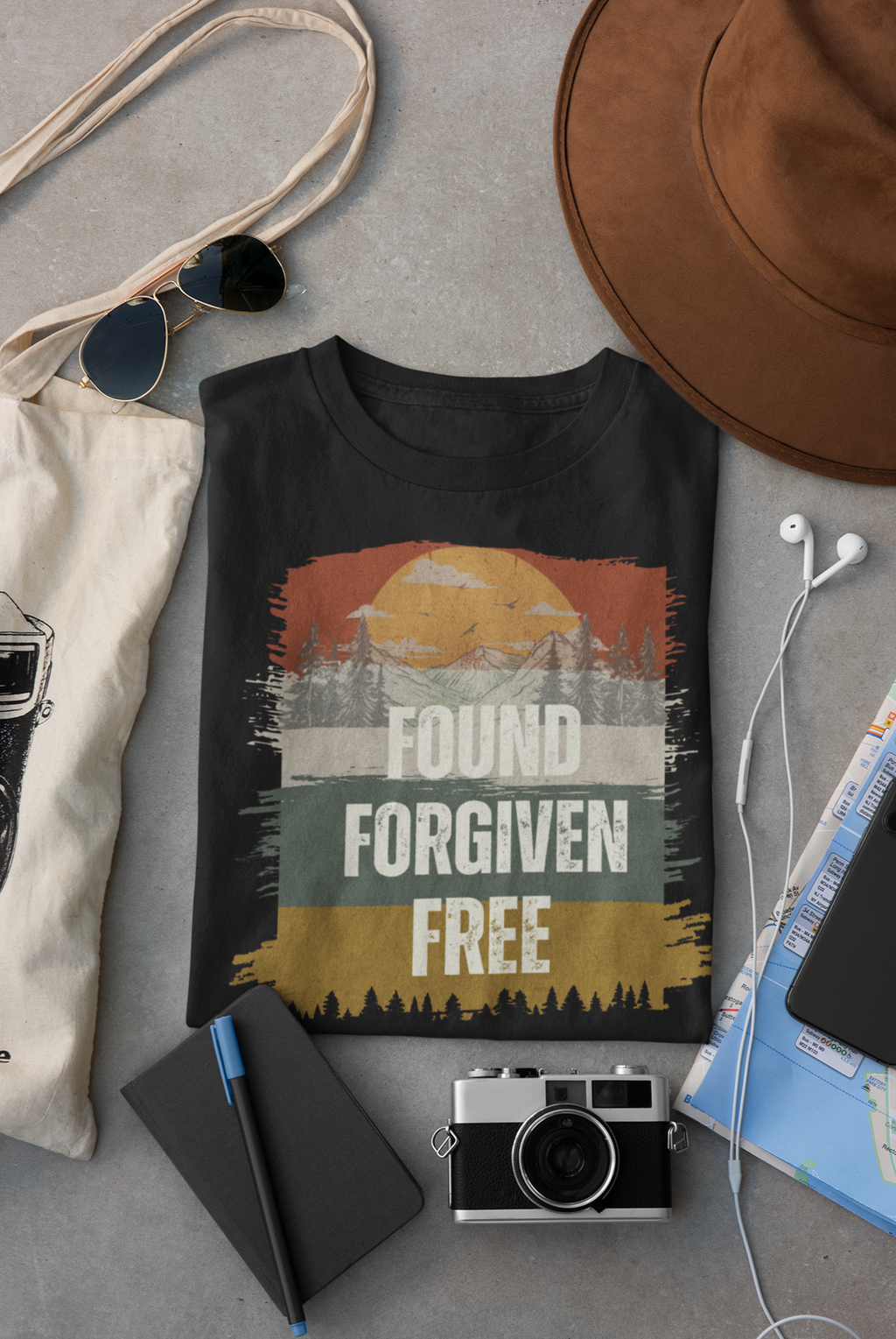 A black Christian graphic T-shirt laid flat on a gray surface, surrounded by lifestyle props including a canvas tote bag, sunglasses, a brown hat, earbuds, a notebook, a camera, and a map. The shirt features a distressed retro sunset graphic with the words 'FOUND FORGIVEN FREE'.