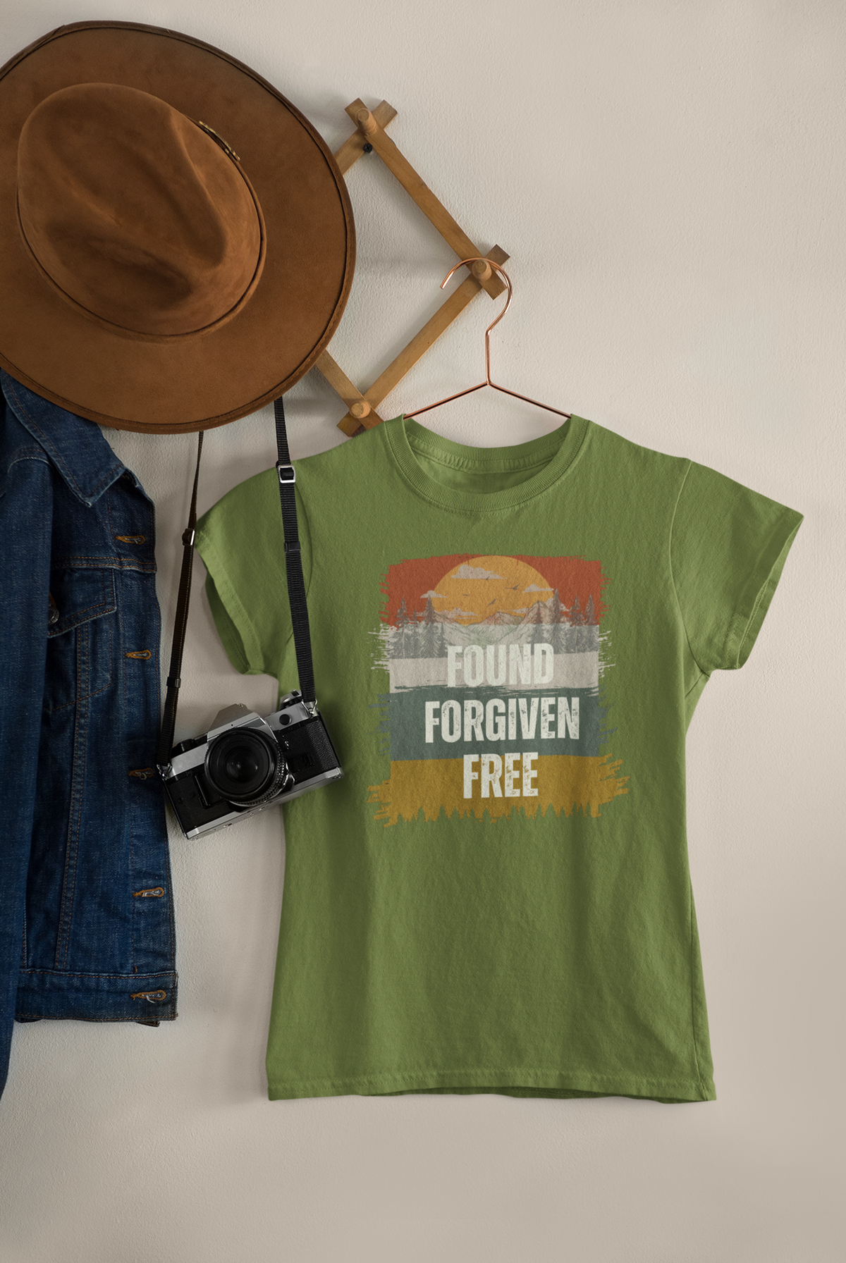 A military green  Christian graphic t-shirt hanging on a wooden hanger against a light beige wall, styled with a brown wide-brim hat, a denim jacket, and a vintage camera.