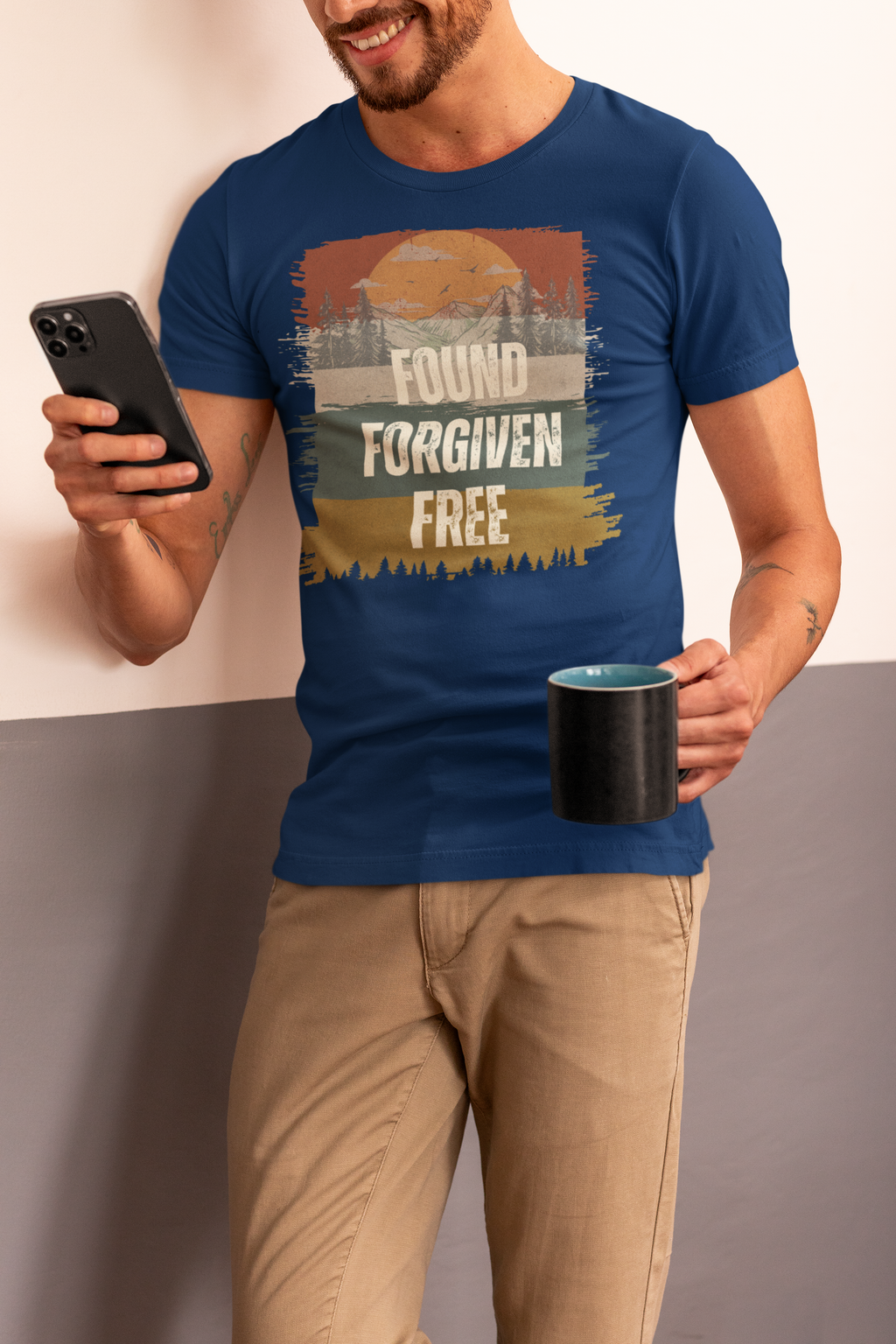 A man wearing a navy blue Christianity graphic t-shirt with a retro sunset design and the text 'FOUND FORGIVEN FREE', paired with khaki pants, holding a black mug and a smartphone.