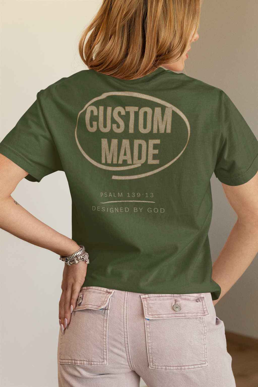 A woman shown from the back wearing an olive green Christian t-shirt with a beige graphic print that reads 'CUSTOM MADE' inside an oval, with smaller text below reading Psalm 139:13/ Designed by God.