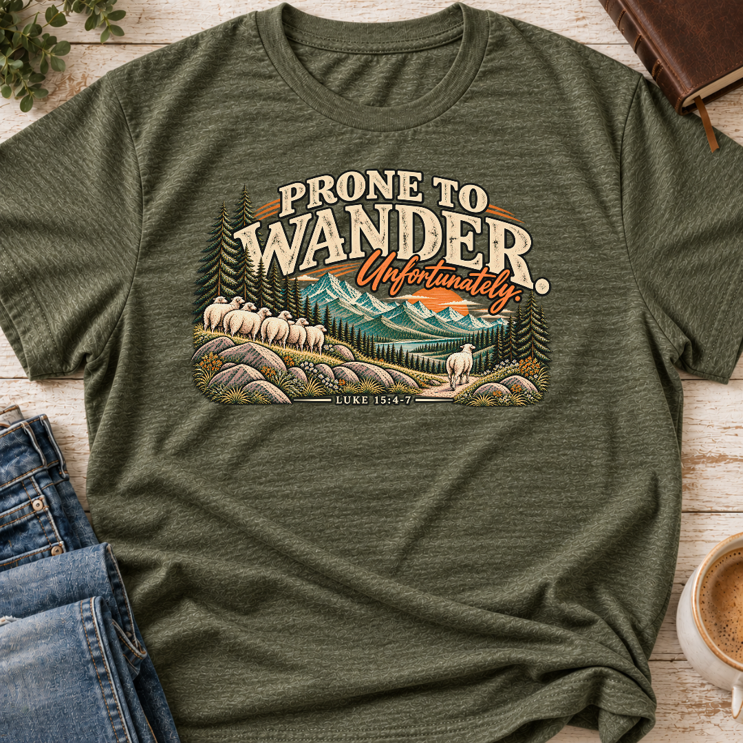 A military green heather Christian graphic t-shirt by SeedSowerGraphics laid flat on a light wood background, styled with blue jeans, a book, and a cup of coffee. The t-shirt features a retro-style scenic graphic with sheep, mountains, and the text 'PRONE TO WANDER.Unfortunately. Luke 15:4-7