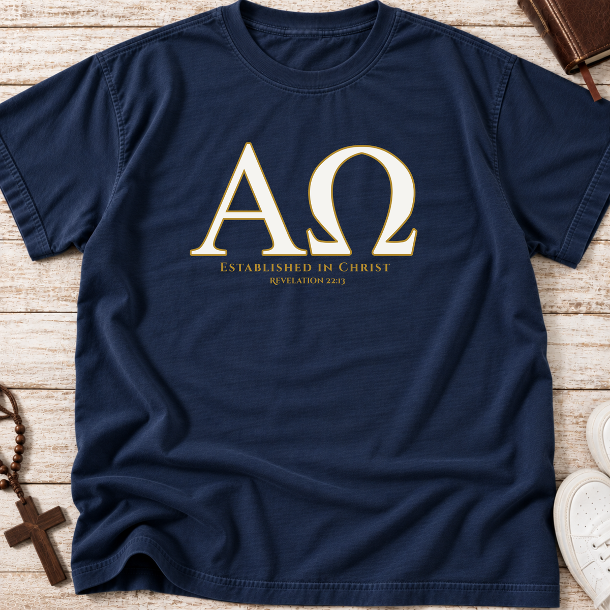 A navy blue short-sleeve t-shirt laid flat on a light wood surface, featuring a large gold Greek letter 'ΑΩ' with the text 'ESTABLISHED IN CHRIST Revelation 22:13' printed below. A brown cross, rosary, book, and white sneakers are partially visible around the edges as styling props.