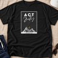 A black short-sleeve graphic T-shirt laid flat on a beige fabric surface, with a white rectangular chest print reading “ACT Justly” above a mountain illustration and “MICAH 6:8.” Blue pants, a white hat, and sunglasses are partially visible as styling props.