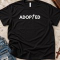 A black short-sleeve t-shirt with the word 'ADOPTED' printed in white across the chest, styled in a flat-lay with blue jeans, a wooden cross, and a leather journal on a textured beige background.