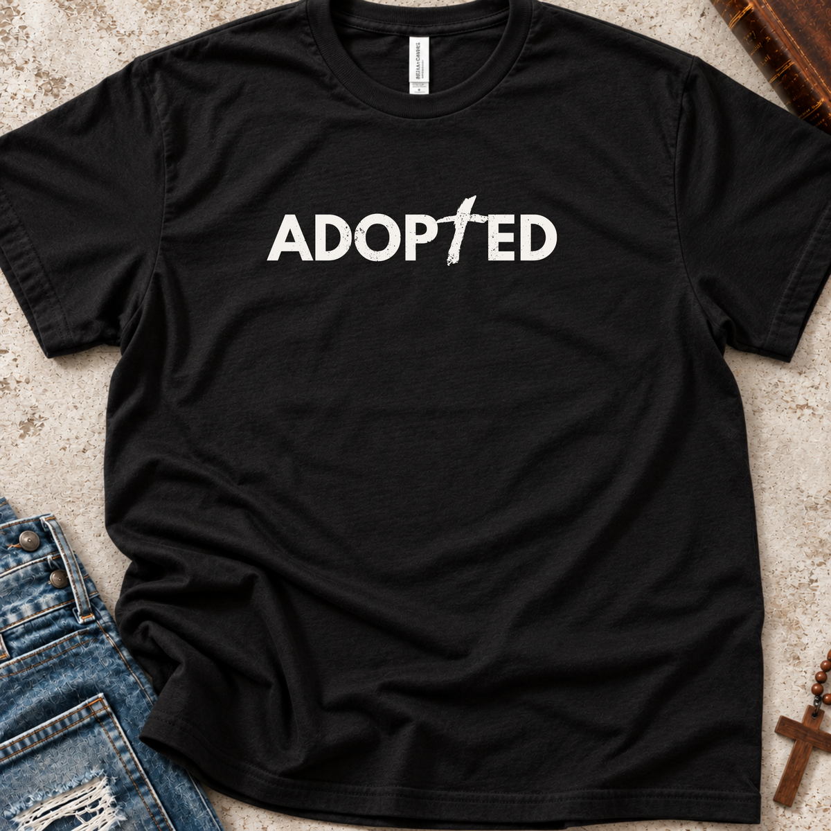 A black short-sleeve t-shirt with the word 'ADOPTED' printed in white across the chest, styled in a flat-lay with blue jeans, a wooden cross, and a leather journal on a textured beige background.