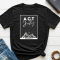 A black short-sleeve graphic T-shirt laid flat on a white wood surface, styled with blue jeans, a closed book, and a beaded bracelet. The shirt has a white rectangular chest graphic reading “ACT Justly” above a mountain illustration with a small cross and “MICAH 6:8” below.