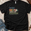 A black short-sleeve graphic T-shirt laid flat on a textured light surface with jeans and a wooden cross as styling props. The shirt has a multicolor typographic slogan reading “BEING NORMAL SEEMS A BIT EXCESSIVE” with “ROMANS 13:2” underneath.