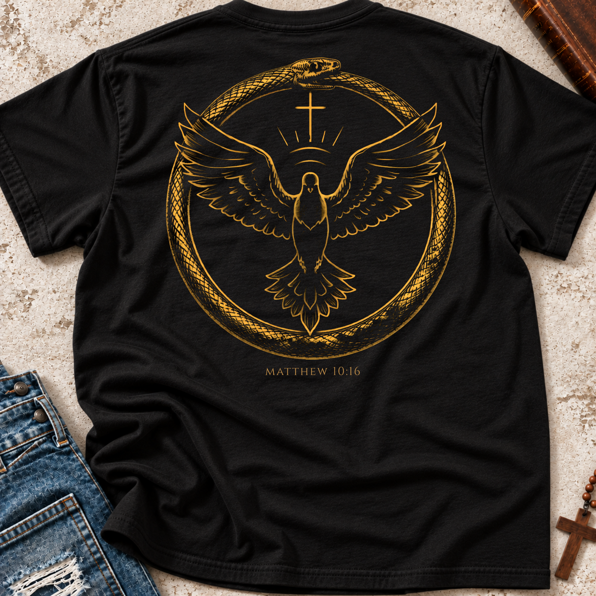 A black short-sleeve T-shirt laid flat on a textured light surface, featuring a large gold graphic on the back with a snake, eagle, cross, and the text 'MATTHEW 10:16'. Blue jeans and small decorative props are partially visible at the edges.