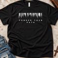 A black short-sleeve t-shirt laid flat on a textured beige surface, featuring a white graphic of a forest silhouette inside a barcode shape above the words 'PONDER YOUR PATH'. Blue jeans are partially visible at the bottom left, and a wooden cross is at the bottom right.