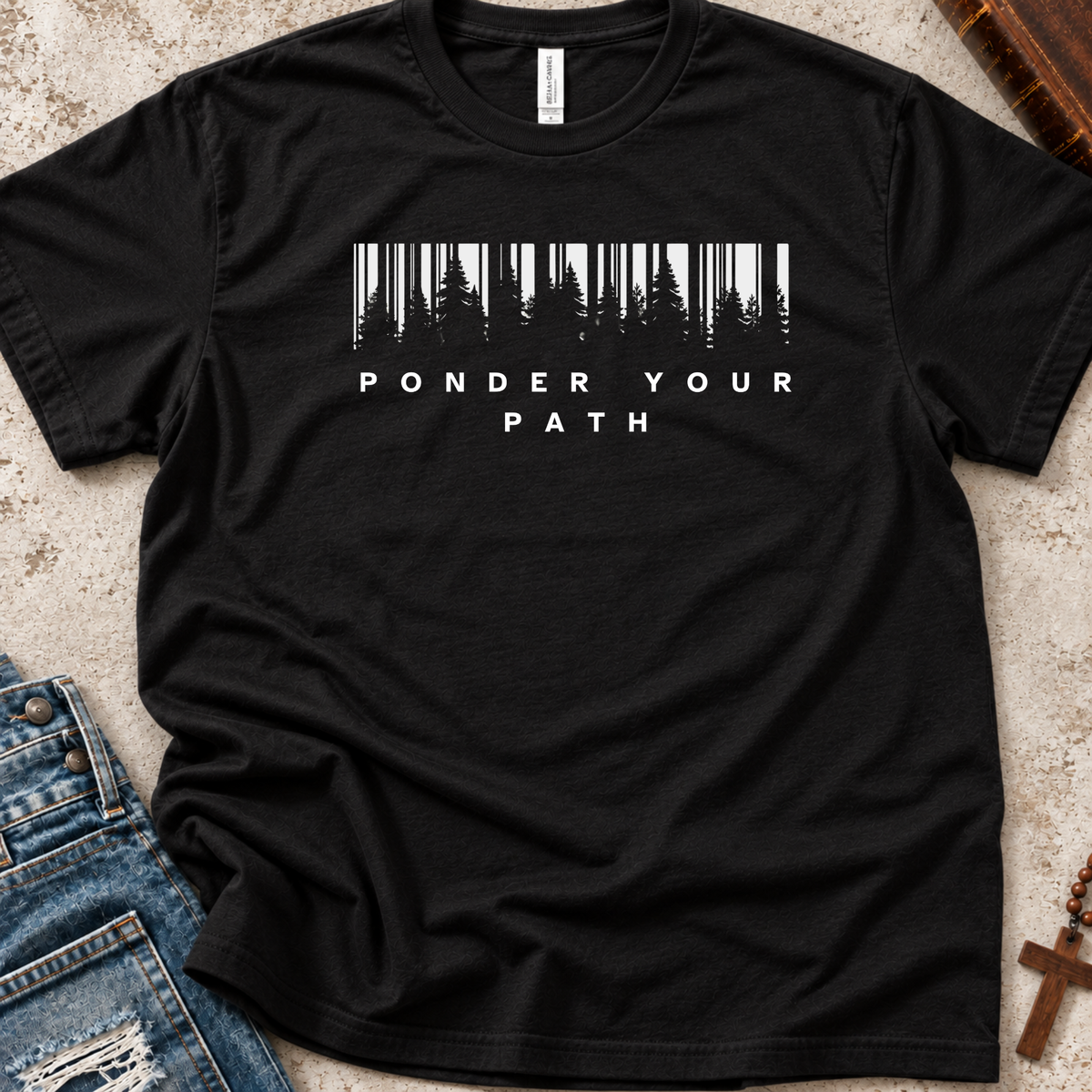 A black short-sleeve t-shirt laid flat on a textured beige surface, featuring a white graphic of a forest silhouette inside a barcode shape above the words 'PONDER YOUR PATH'. Blue jeans are partially visible at the bottom left, and a wooden cross is at the bottom right.