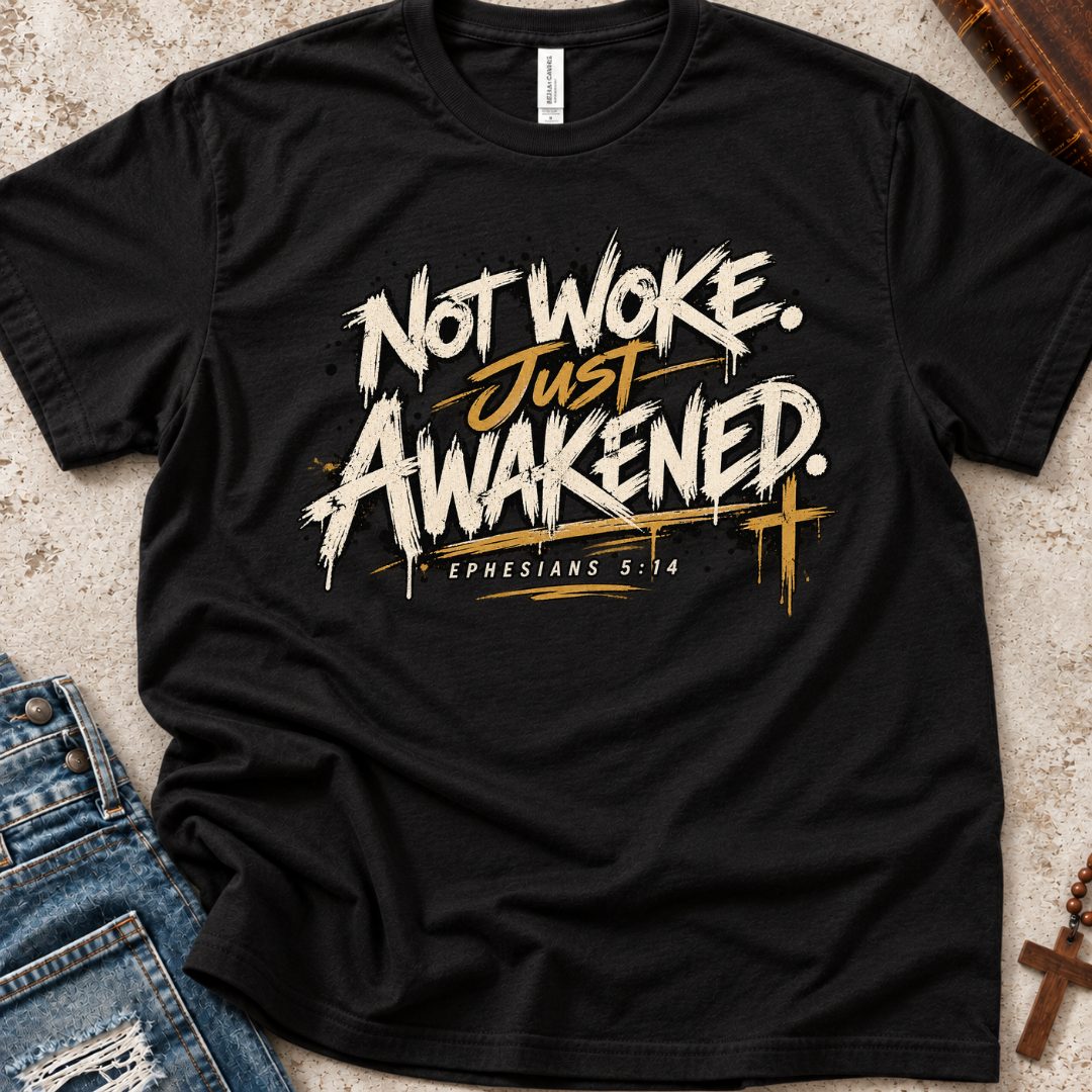 A black short-sleeve Christian graphic T-shirt by SeedSowerGraphics laid flat on a textured light surface, styled with blue jeans, a wooden cross, and a rosary. The shirt features the phrase 'NOT WOKE. Just Awakened. Ephesians 5:14