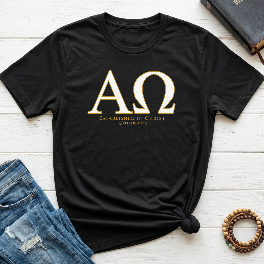 A black short-sleeve T-shirt laid flat on a white wood surface, styled with blue jeans, a book, and a beaded bracelet. The shirt has large gold Greek letters with smaller gold text below reading 'ESTABLISHED IN CHRIST' and 'REVELATION 22:13'.