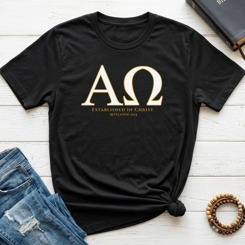 A black short-sleeve T-shirt laid flat on a white wood surface, styled with blue jeans, a book, and a beaded bracelet. The shirt has large gold Greek letters with smaller gold text below reading 'ESTABLISHED IN CHRIST' and 'REVELATION 22:13'.