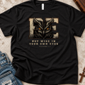A black short-sleeve graphic T-shirt laid flat on a textured light surface with jeans and a wooden cross as styling props. The shirt features a large distressed beige and brown letter E with an owl face inside it, orange eyes, and the text 'NOT WISE IN YOUR OWN EYES' and 'PROVERBS 3:7' below.