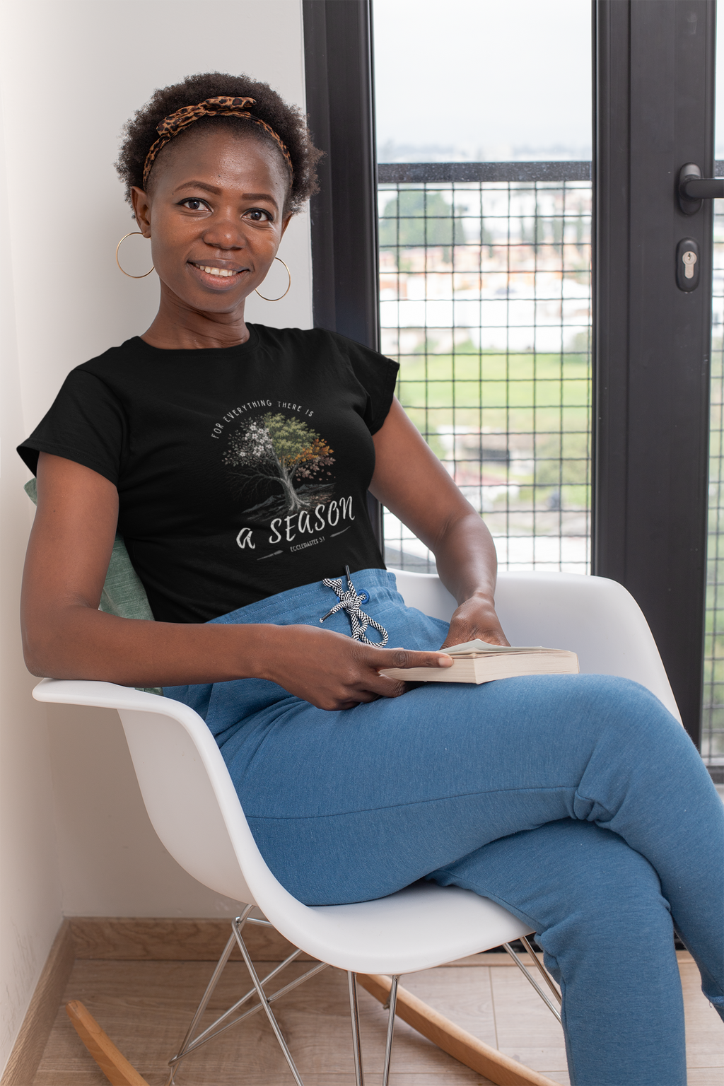 A woman sitting in a white rocking chair by a window, wearing a black Christian graphic t-shirt by SeedSowerGraphics that reads 'For Everything There is a Season' with a tree illustration and blue jeans.