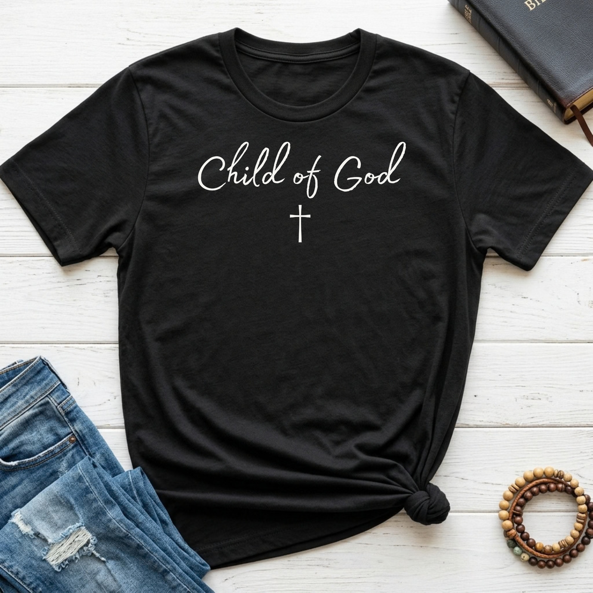 A black short-sleeve t-shirt with 'Child of God' and a cross printed in white on the chest, styled in a flat lay with blue jeans, a leather Bible, and wooden beads on a white wood plank background.