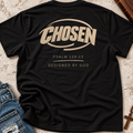 A black short-sleeve t-shirt laid flat on a textured light beige surface, featuring a large beige graphic on the back that reads 'CHOSEN' with 'PSALM 139:13' and 'DESIGNED BY GOD' below. Blue jeans and a wooden cross are partially visible as styling props.