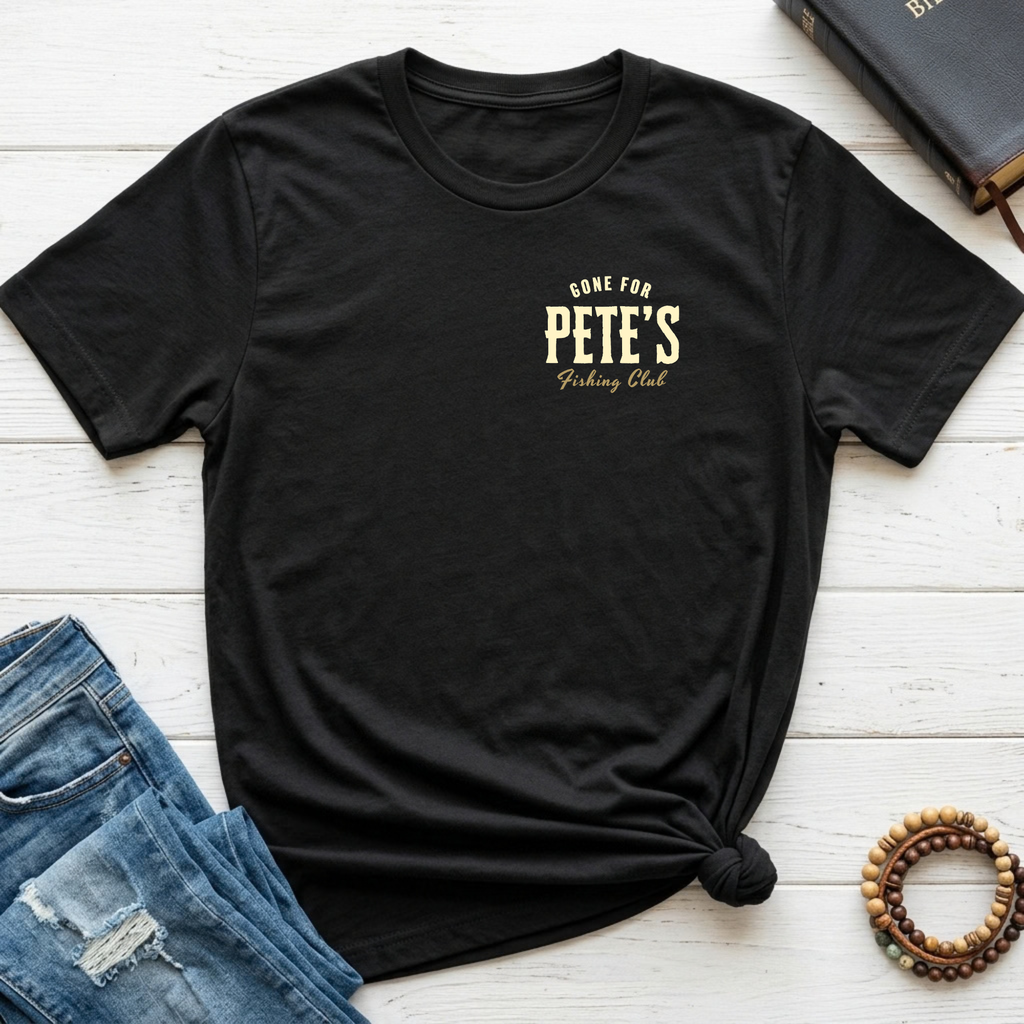 A black short-sleeve graphic T-shirt laid flat on a white wood surface, styled with blue jeans, a book, and a beaded bracelet. The shirt has a small gold chest print reading 'Gone For PETE'S Fishing Club' and is tied at the hem.