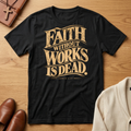 A black short-sleeve graphic T-shirt laid flat on a wooden surface, featuring the phrase 'FAITH WITHOUT WORKS IS DEAD.' in large distressed gold lettering with 'JAMES 2:17' printed below.