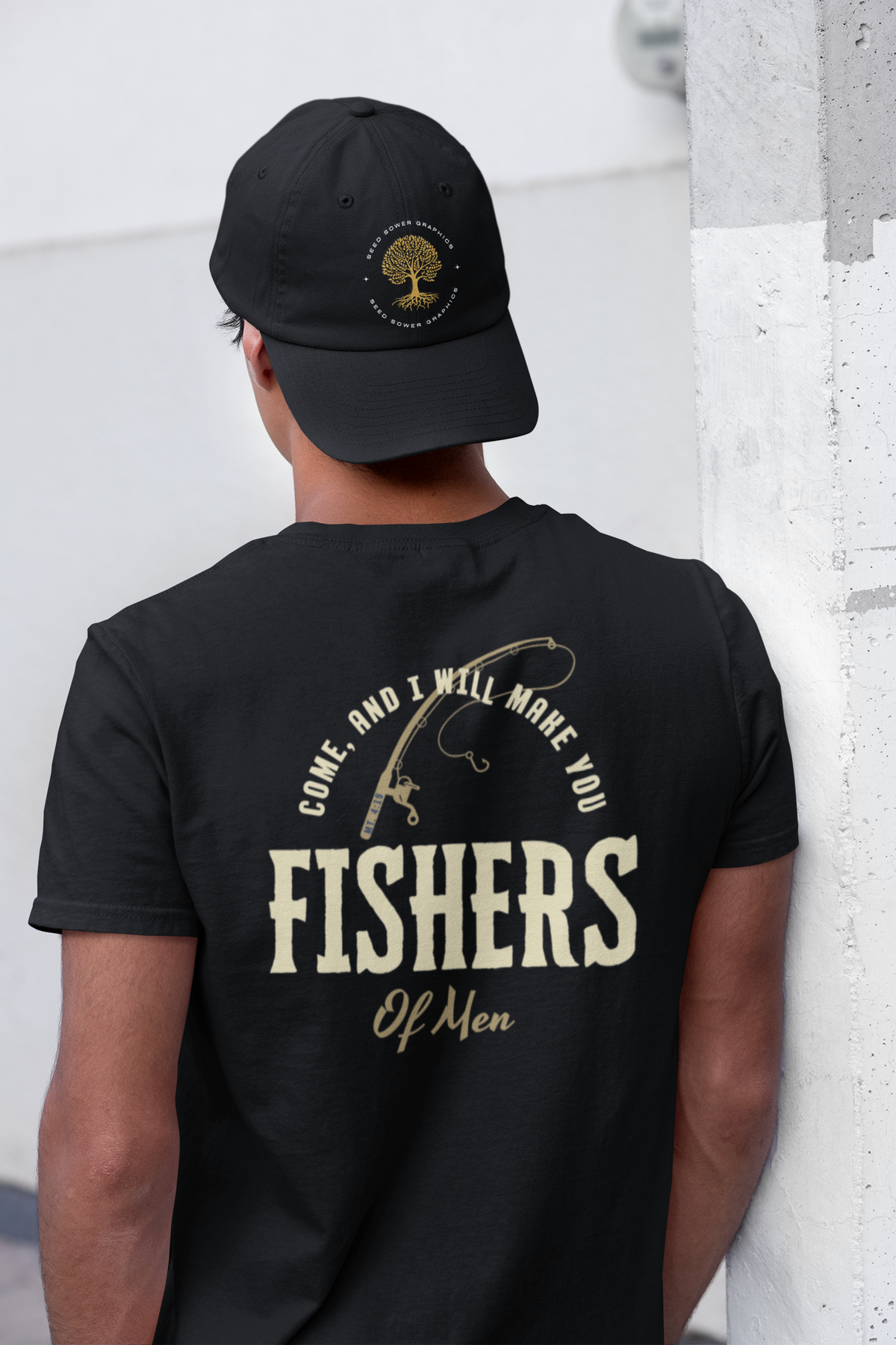 A man viewed from behind wearing a black t-shirt and a black baseball cap. The back of the t-shirt features a cream-colored graphic with a fishing rod, the text 'COME, AND I WILL MORE YOU FISHERS Of Men', and a circular tree logo on the cap.