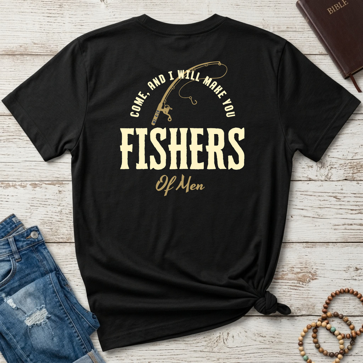 A black short-sleeve Christian graphic T-shirt laid flat on a whitewashed wood surface, styled with jeans, a brown leather Bible, and wooden beads. The shirt has a large cream graphic on the back featuring a fishing rod and the text “COME, AND I WILL MAKE YOU FISHERS OF MEN.