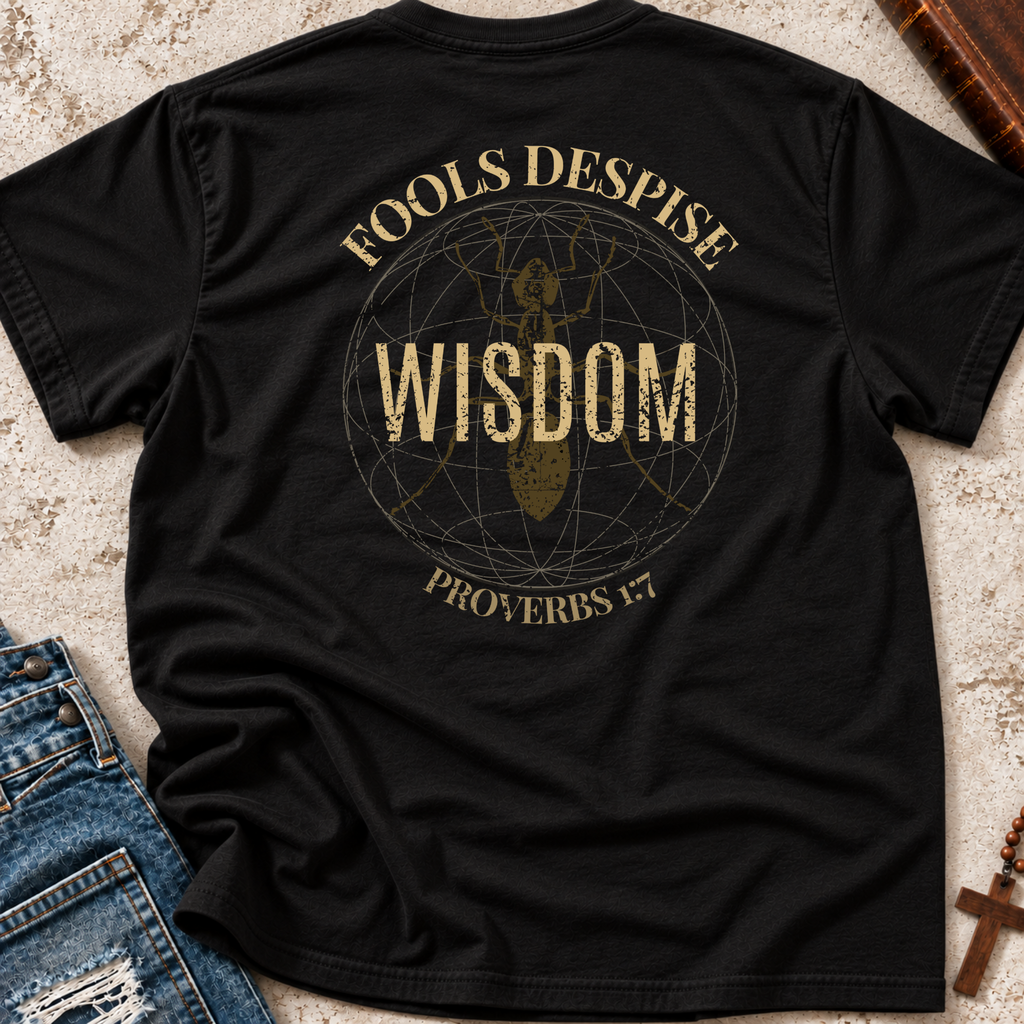 A black short-sleeve graphic T-shirt laid flat on a textured beige surface, with blue jeans partly visible at the lower left and a wooden book or box at the upper right. The shirt features a distressed gold circular design with the words 'FOOLS DESPISE WISDOM' and 'PROVERBS 1:7' around a central figure.
