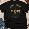 A black short-sleeve graphic T-shirt laid flat on a textured beige surface, with blue jeans partly visible at the lower left and a wooden book or box at the upper right. The shirt features a distressed gold circular design with the words 'FOOLS DESPISE WISDOM' and 'PROVERBS 1:7' around a central figure.