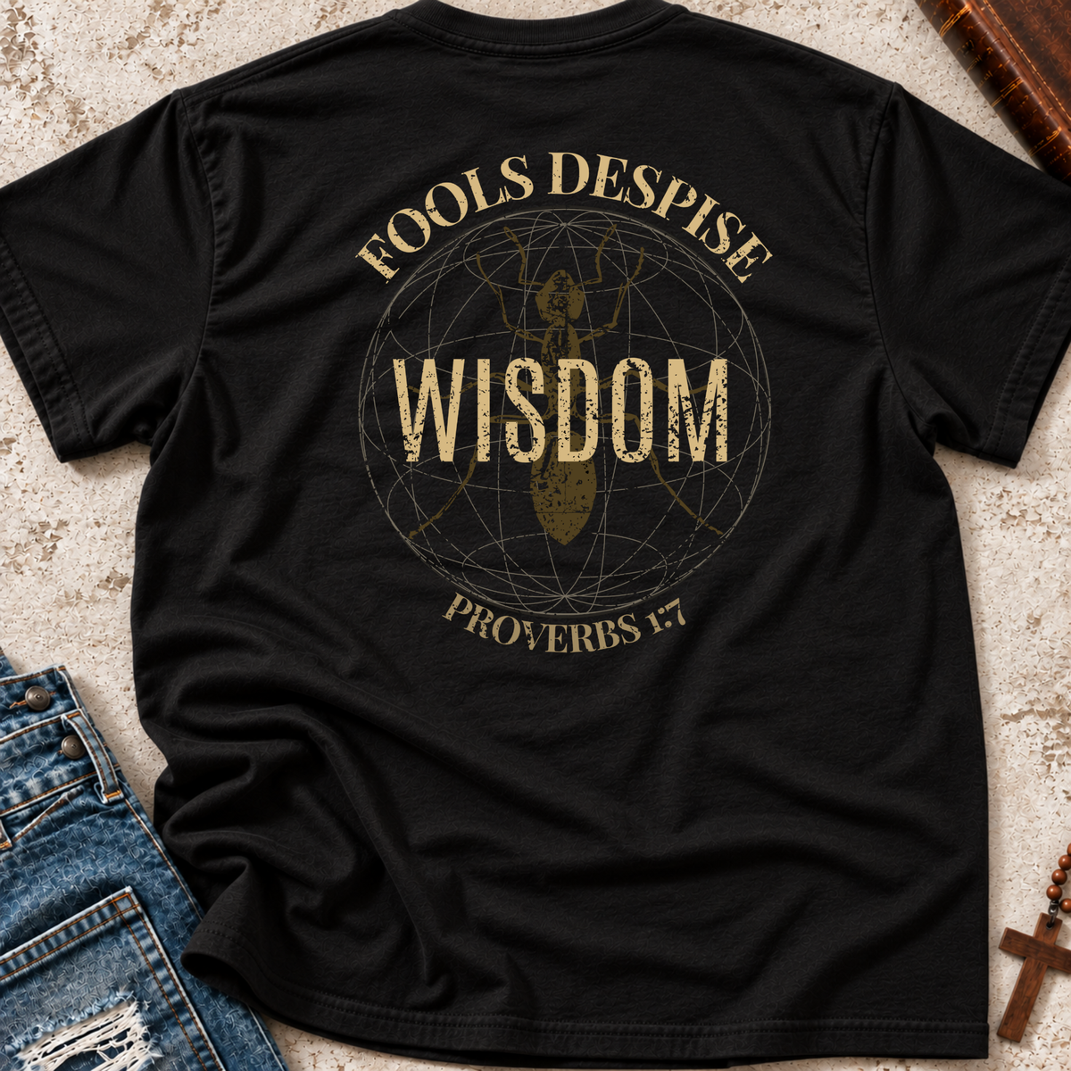 A black short-sleeve graphic T-shirt laid flat on a textured beige surface, with blue jeans partly visible at the lower left and a wooden book or box at the upper right. The shirt features a distressed gold circular design with the words 'FOOLS DESPISE WISDOM' and 'PROVERBS 1:7' around a central figure.