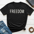 A black short-sleeve graphic T-shirt laid flat on a white wood surface, styled with blue jeans, a book, and a beaded bracelet. The shirt has large white text reading 'STRENGTH IN WEAKNESS' with smaller text '2 CORINTHIANS 12:9' underneath.