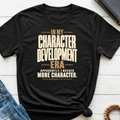 A black Christian graphic t-shirt by SeedSowerGraphics with a warm beige text print reading 'IN MY CHARACTER DEVELOPMENT ERA APPARENTLY, I NEEDED MORE CHARACTER. ROMANS 5:3-4'.