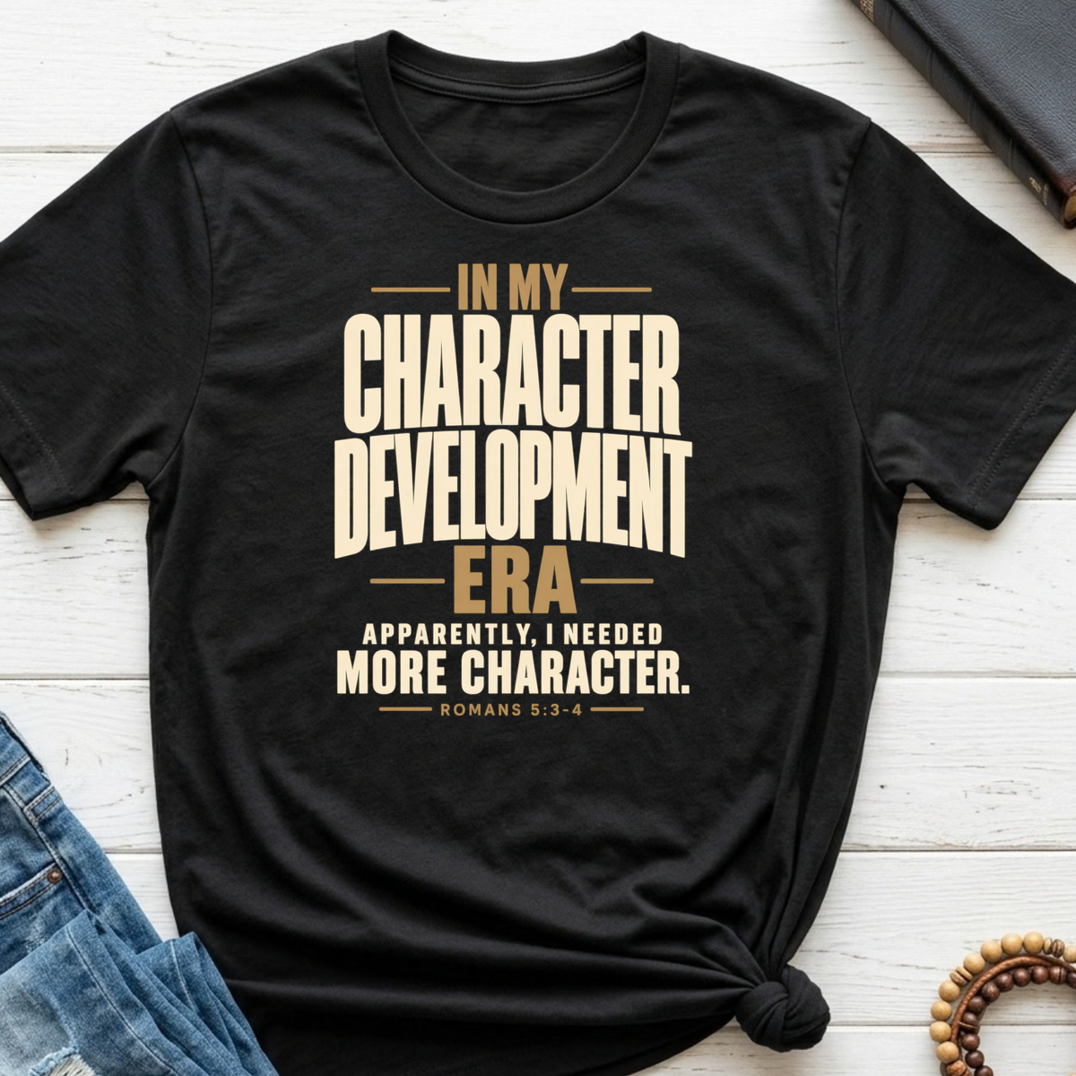 A black Christian graphic t-shirt by SeedSowerGraphics with a warm beige text print reading 'IN MY CHARACTER DEVELOPMENT ERA APPARENTLY, I NEEDED MORE CHARACTER. ROMANS 5:3-4'.