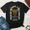 A black short-sleeve Christian graphic T-shirt by SeedSowerGraphics laid flat on a whitewashed wood surface, styled with blue jeans, a brown leather Bible, and a beaded bracelet. The shirt features a large distressed gold eagle graphic with white text reading 'SOAR ON WINGS LIKE EAGLES' and 'Isaiah 40:31' printed below.