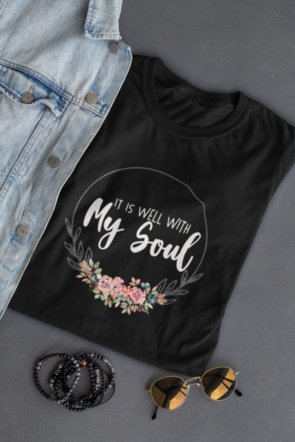 A black Christian graphic t-shirt by SeedSowerGraphics with a floral wreath design and the text 'IT IS WELL WITH My Soul' in white script, styled in a flat lay with a denim jacket, beaded bracelets, and sunglasses on a gray background.