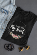 A black Christian graphic t-shirt by SeedSowerGraphics with a floral wreath design and the text 'IT IS WELL WITH My Soul' in white script, styled in a flat lay with a denim jacket, beaded bracelets, and sunglasses on a gray background.