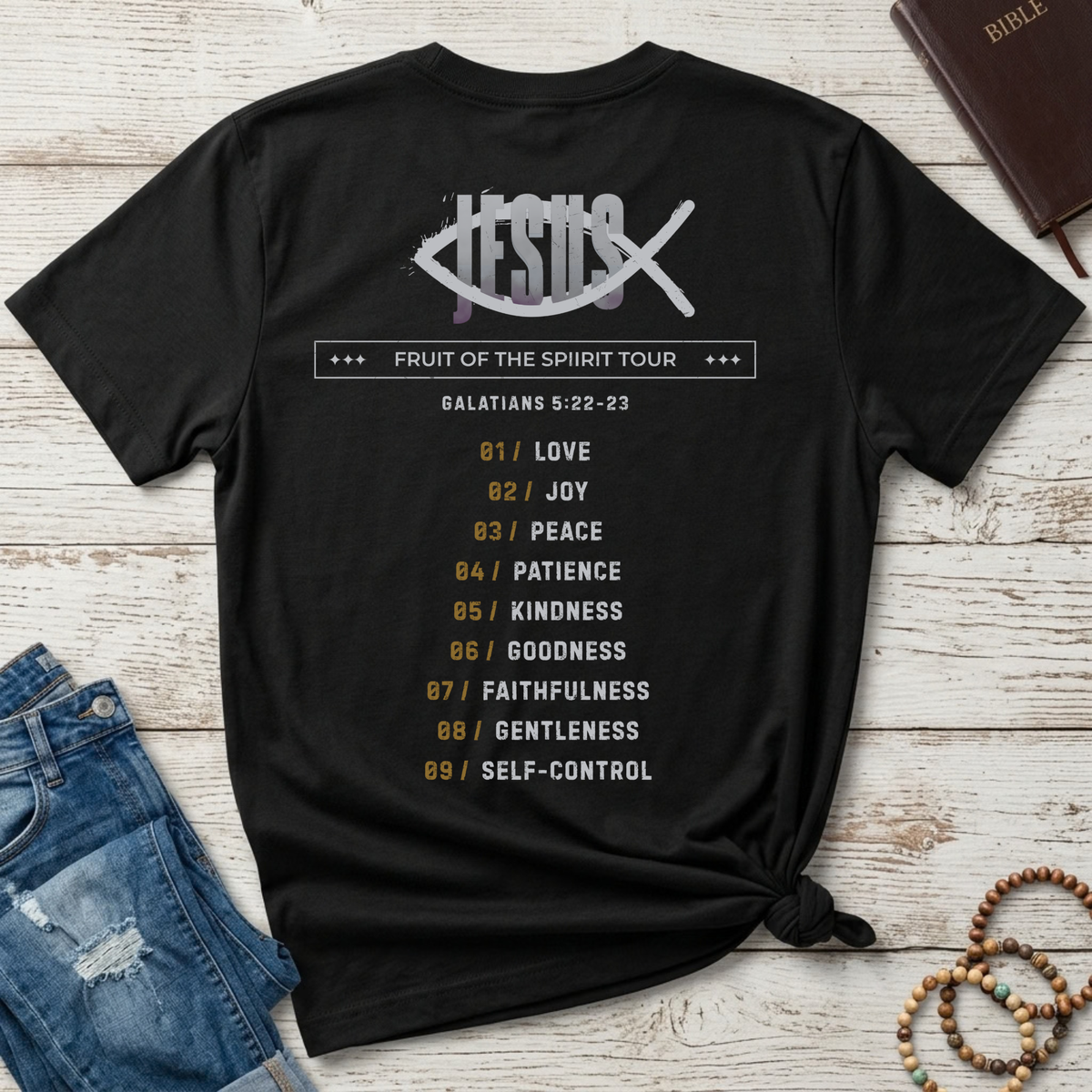 A black Christian graphic t-shirt by SeedSowerGraphics laid flat on a whitewashed wood background, styled with blue jeans, a brown leather Bible, and wooden beads. The back of the shirt features a white fish logo with 'JESUS' crossed through it, the text 'FRUIT OF THE SPIRIT TOUR', and a numbered list from 01 to 09 in gold and white.