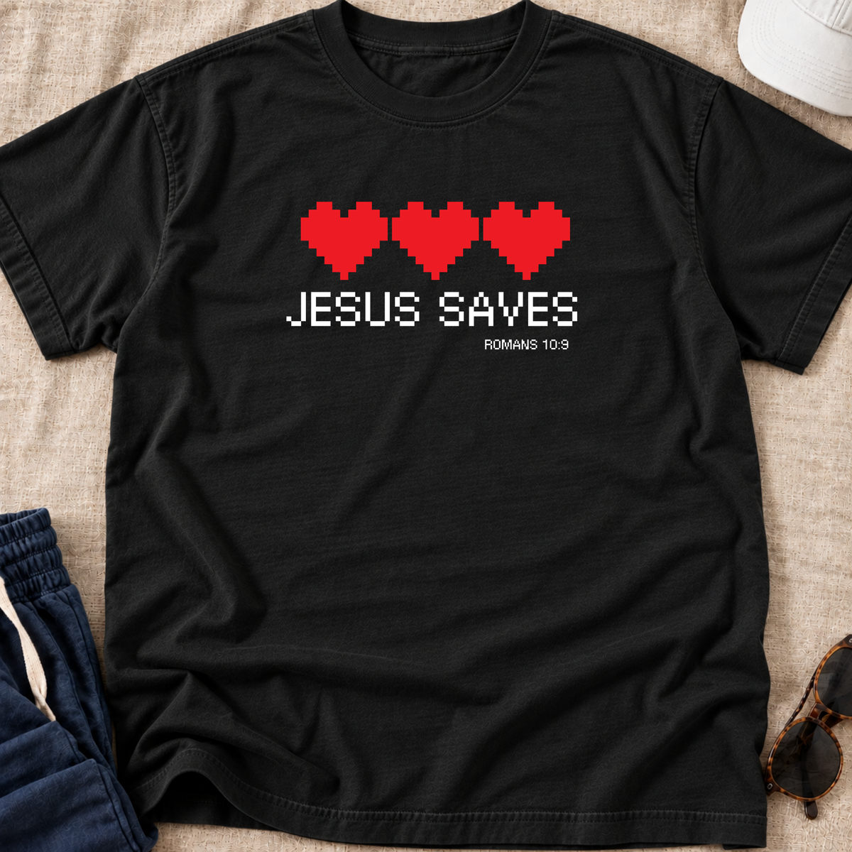 A black short-sleeve t-shirt laid flat on a beige fabric surface, featuring four red pixel hearts and the text 'JESUS SAVES' and 'ROMANS 10:9' in white pixel-style lettering. Partial jeans, sunglasses, and a hat are visible around the edges as styling props.