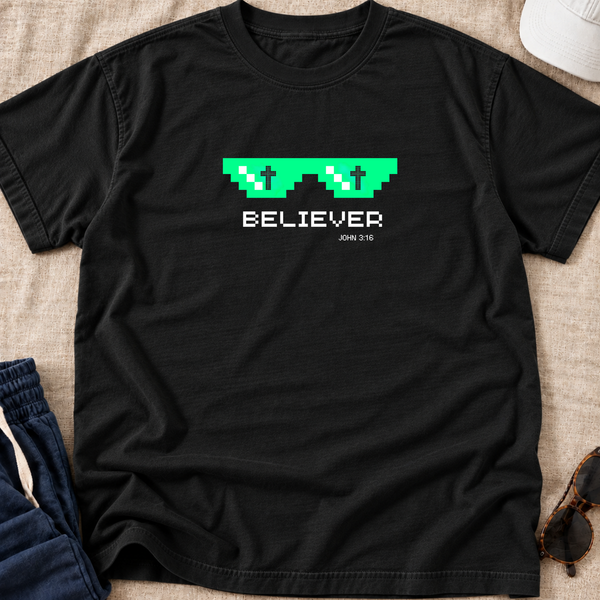 A black short-sleeve T-shirt laid flat on a beige fabric surface, featuring a green pixelated sunglasses graphic with white crosses inside and the word 'BELIEVER' in white below, with 'JON 3:16' printed underneath. Partially visible props include blue jeans, a white cap, and sunglasses.