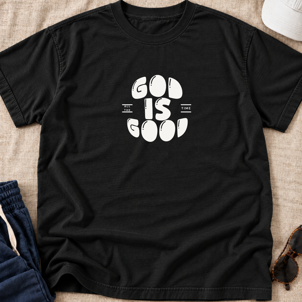 A black short-sleeve t-shirt with a white graphic print reading 'GOD IS GOOD' laid flat on a beige textured fabric background, styled with blue jeans, a white cap, and sunglasses.