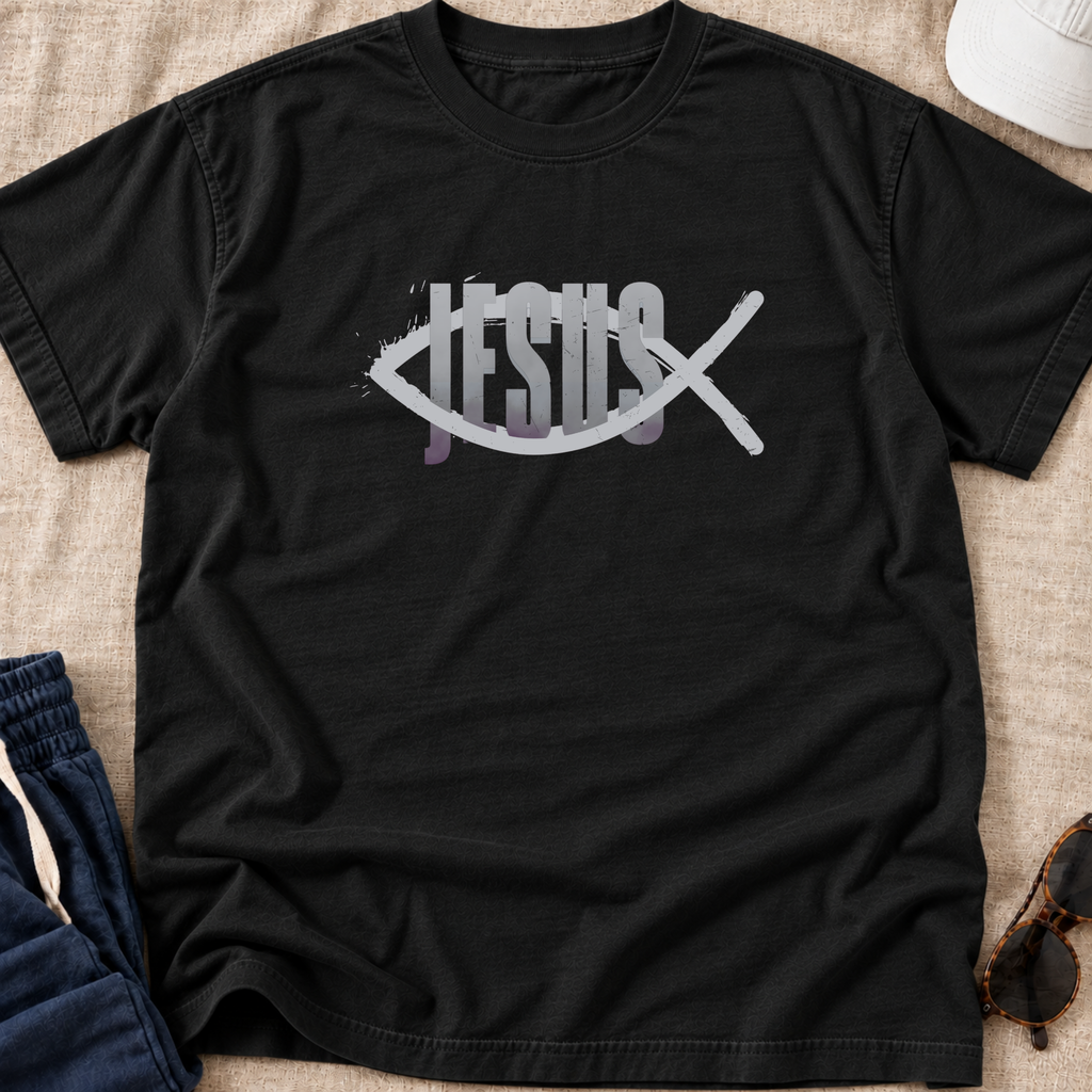 A black short-sleeve Christian graphic t-shirt laid flat on a beige fabric surface, featuring a white fish symbol with the word 'JESUS' integrated through it and a white diagonal cross over the design. Partially visible props include blue pants, sunglasses, and a white cap.
