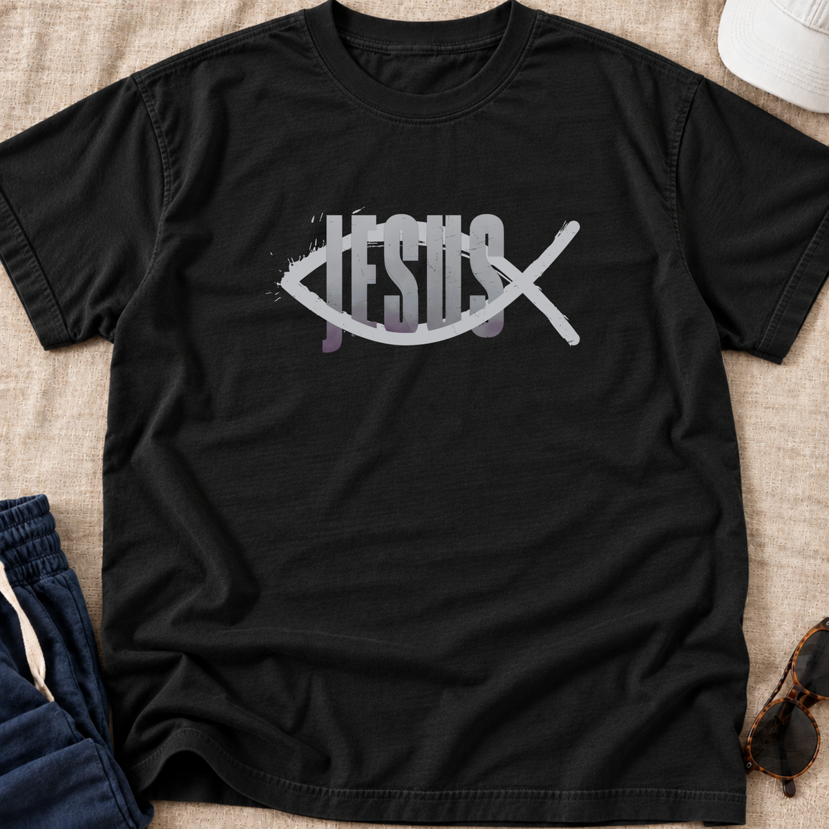 A black short-sleeve Christian graphic t-shirt laid flat on a beige fabric surface, featuring a white fish symbol with the word 'JESUS' integrated through it and a white diagonal cross over the design. Partially visible props include blue pants, sunglasses, and a white cap.