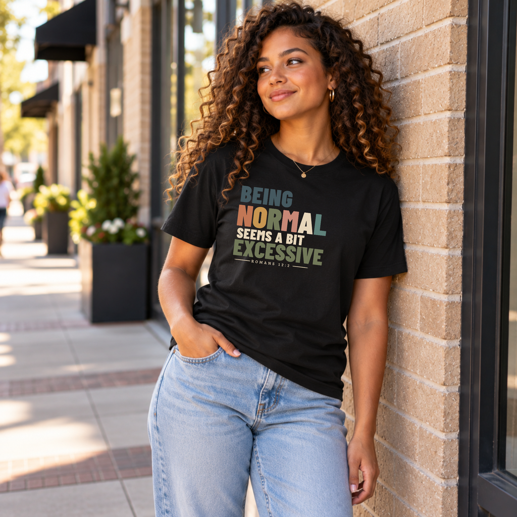 A woman with curly hair leans against a brick wall on a sidewalk, wearing a black graphic T-shirt that reads 'BEING NORMAL SEEMS A BIT EXCESSIVE' in multicolor text, paired with light blue jeans.