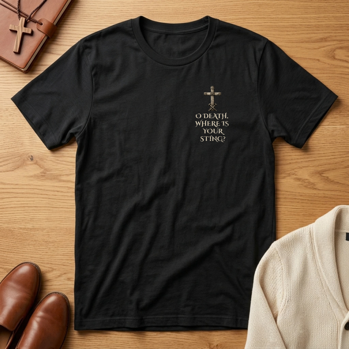 A black short-sleeve crewneck t-shirt with a small gold cross graphic and the text 'O DEATH WHERE IS YOUR STING?' on the left chest, presented in a flat-lay arrangement on a wooden table with a brown leather wallet, brown shoes, and a cream cardigan.
