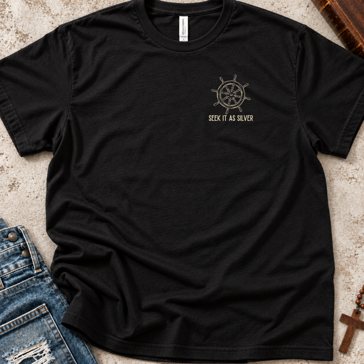 A black short-sleeve t-shirt with a small beige graphic of a ship's wheel and the text 'SEEK IT AS SILVER' on the left chest, styled flat-lay on a textured surface with jeans and wooden props.