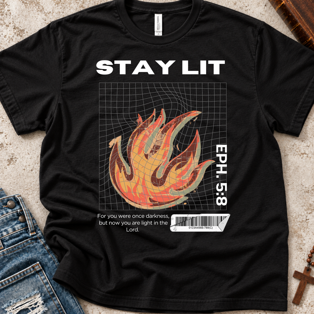 A black graphic t-shirt laid flat on a textured beige surface with denim jeans and a wooden cross as styling props. The shirt features a white 'STAY LIT' text at the top, a central flame graphic in orange, yellow, and red over a grid, the text 'EPH. 5:8'