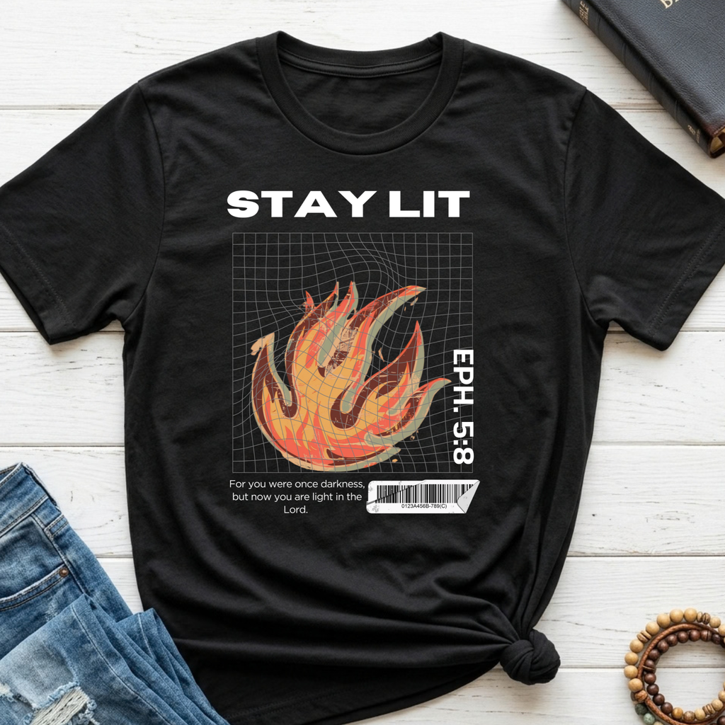 A black graphic t-shirt laid flat on a white wood background, styled with blue jeans, a book, and wooden beads. The t-shirt features a white 'STAY LIT' text at the top, a 3D wireframe flame graphic in orange and yellow, and a Bible verse from Ephesians 5:8 with a barcode graphic.